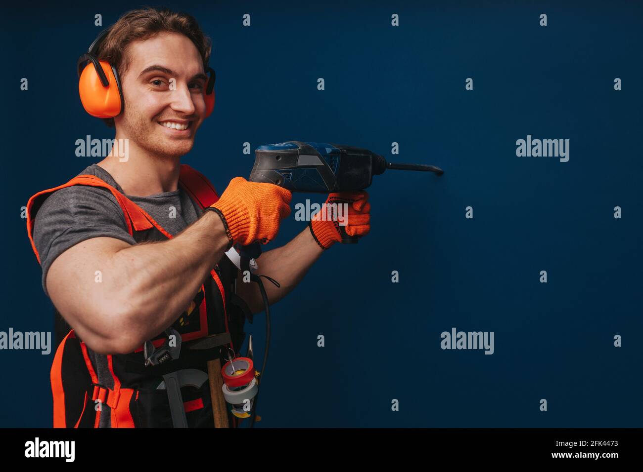 Young builder is drilling the wall and smiling, isolated over blue background Stock Photo