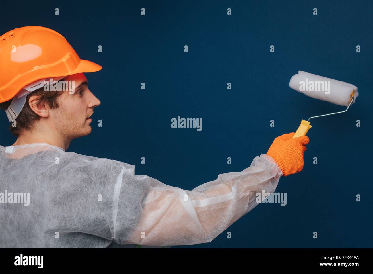 Builder is using roller to paint wall, isolated over blue background Stock Photo
