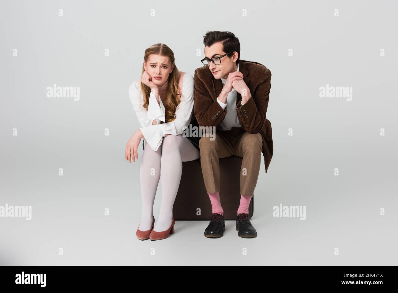 upset couple in vintage outfit sitting on suitcase on grey background Stock Photo