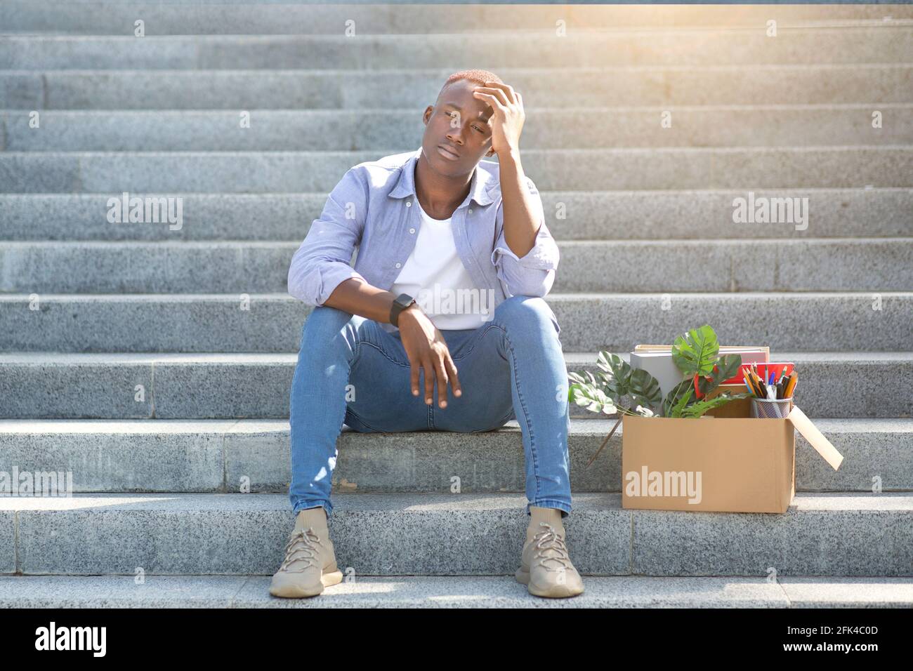 Crisis and unemployment. Stressed black guy with personal stuff sitting on stairs after losing his job, outdoors Stock Photo