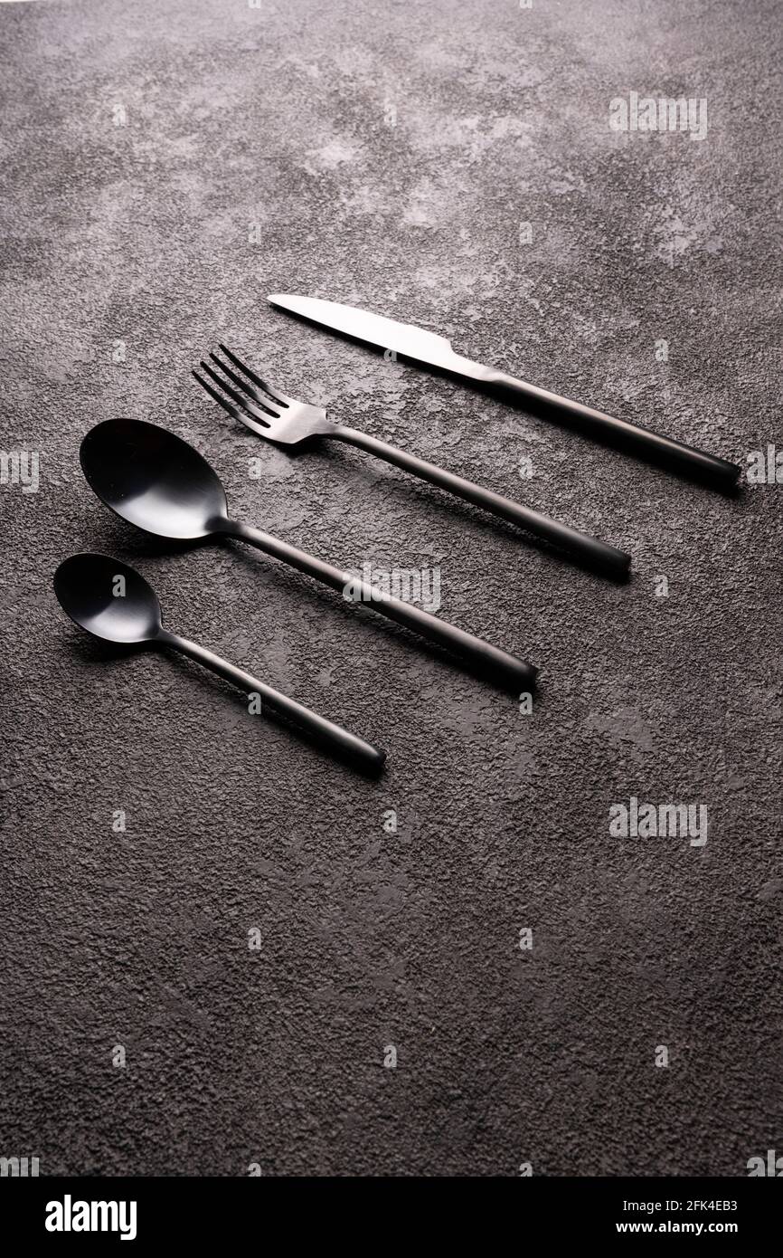 Black cutlery-fork, spoon, knife, on a dark background in the center. Minimalistic still life, vertical photo. Stock Photo