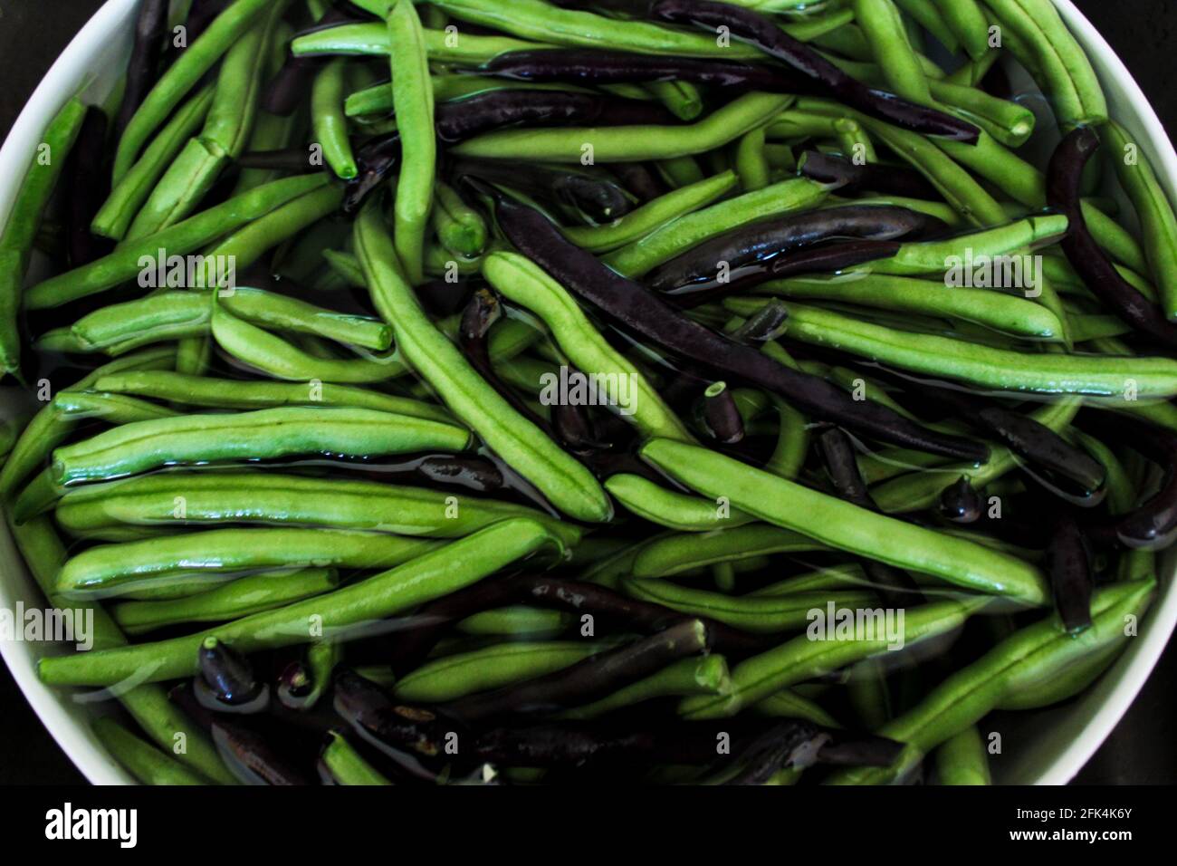 many green and purple beans immersed in water in a white bowl Stock Photo