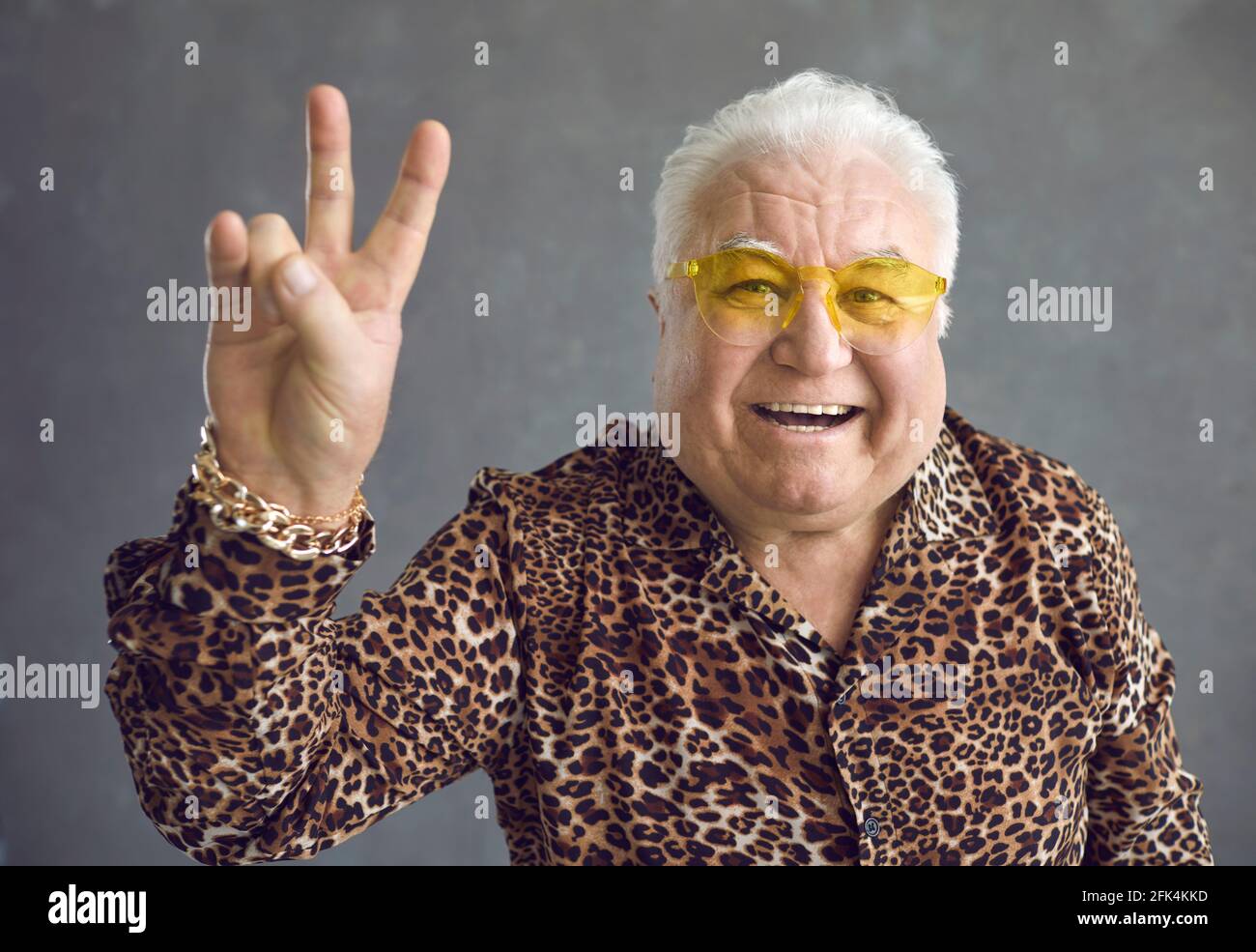 Cheerful stylish glamorous senior man showing v-sign peace symbol on gray background. Stock Photo