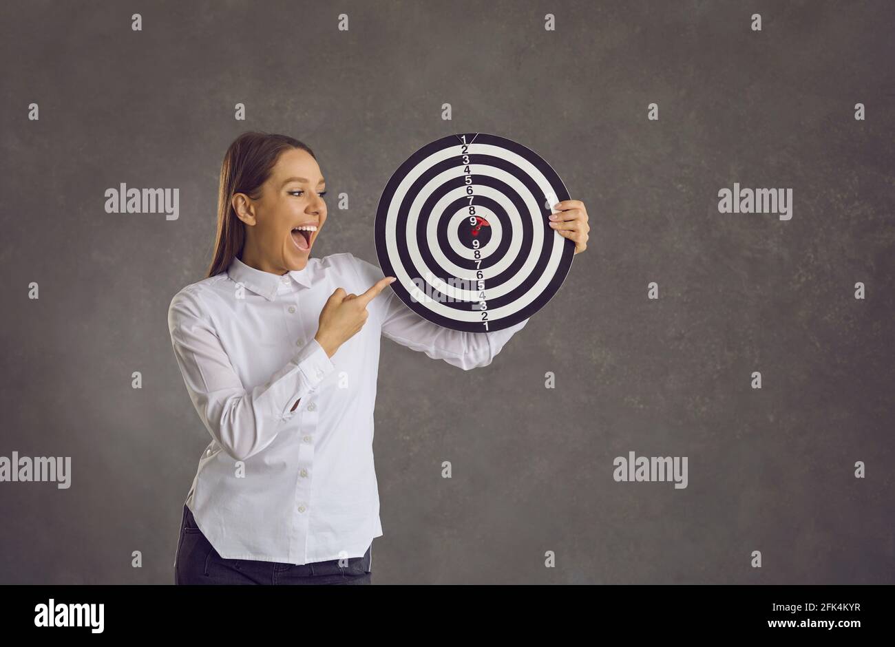 Happy young businesswoman holding dartboard target with red dart right in bullseye Stock Photo