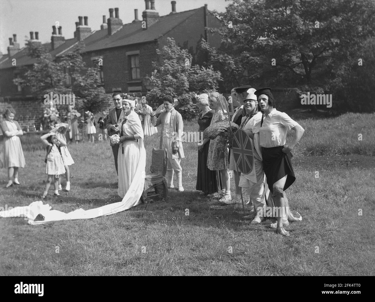 1950s, historical, on a piece of land at the end of terrace of victorian houses, a group of local men - 'the Heavy Gang' dressed in womens clothes and fancy dress standing for a photo before a performance for the 'May Day' festival, England, UK. The amateur actors here are in differen costumes, including Guinevere, the legendary Queen of King Arthur. May Day was an ancient tradition which celebrated the arrival of Spring and Summer and performers would  dress up and re-enact old folklore and stories with dancing around a tall wooden pole, a maypole, a popular activity. Stock Photo