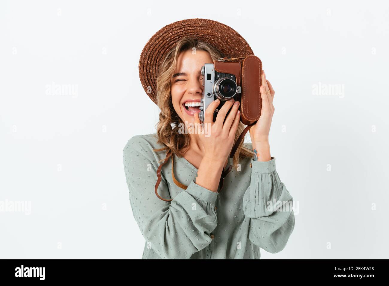 Joyful young woman wearing summer hat makes photos while looking through the viewfinder of her vintage camera, white background Stock Photo