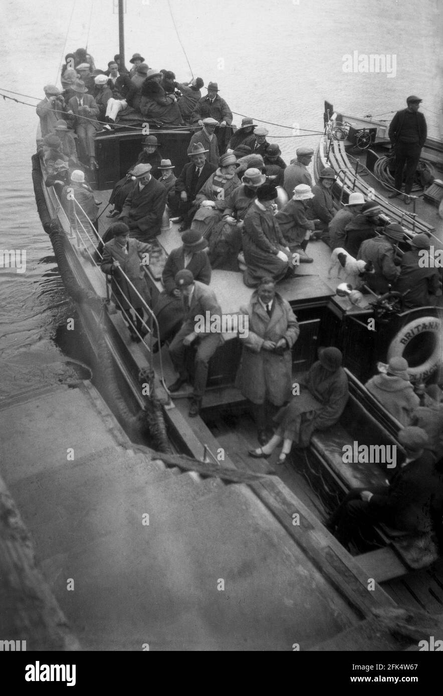 1930s, Beside some steps, at the quayside, possibly at Cleethorpes, flat caps a plenty as men, women and school children crowd on the pleasure boat, 'Britannia' for a trip. An excursion by boat from Bridlington to the East Yorkshire seaside resort was popular in this era and up to the mid- 1950s, there were five large pleasure boats offering such journeys. Built in 1923 by Hayward & Croxan in Southend-on-Sea, the motor boat, Britannia ran excursions at Bridlington until the late 1960s. She was an open boat of between 50ft and 60ft and passengers sat where they could find a space. Stock Photo