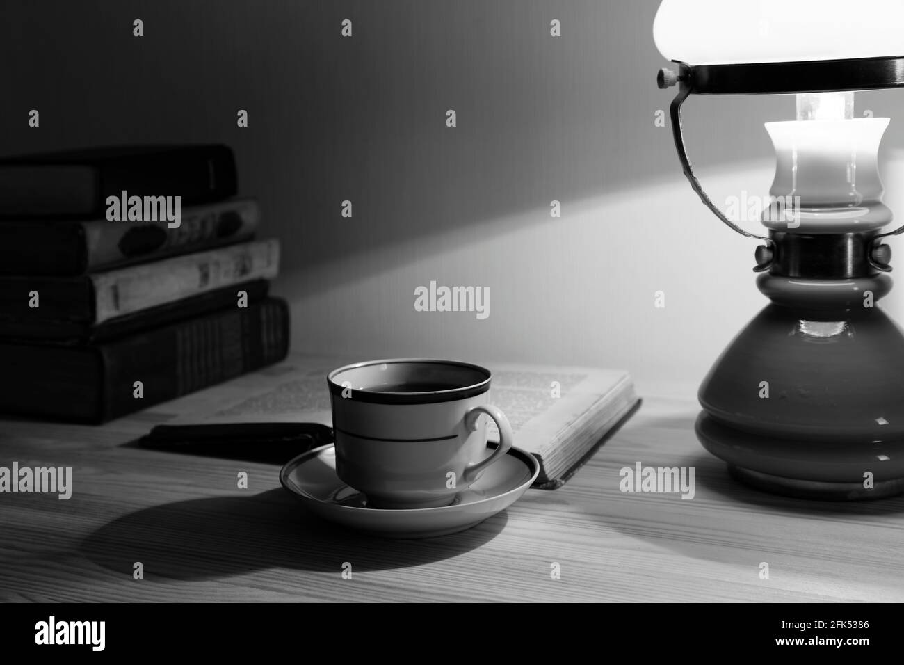 Cup of tea and stack of old books on the table with lamp. Black and white. Stock Photo