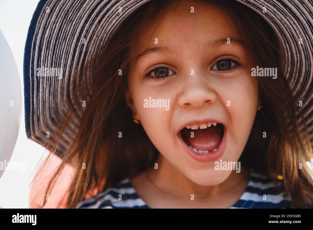 Close up fun portrait of cute laughing little girl in striped sunhat and shirt looking at camera. Selective focus Stock Photo