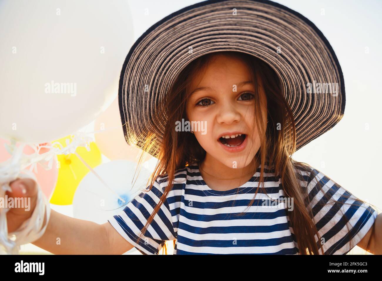 Close up fun portrait of cute laughing little girl holding colorful balloons in striped sunhat and shirt looking at camera Stock Photo