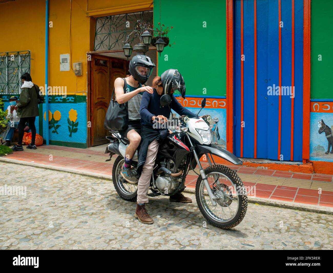 Guatapé, Antioquia, Colombia - April 4 2021: Woman Riding a Motorcycle and a Young White Man Getting on Behind Her Stock Photo