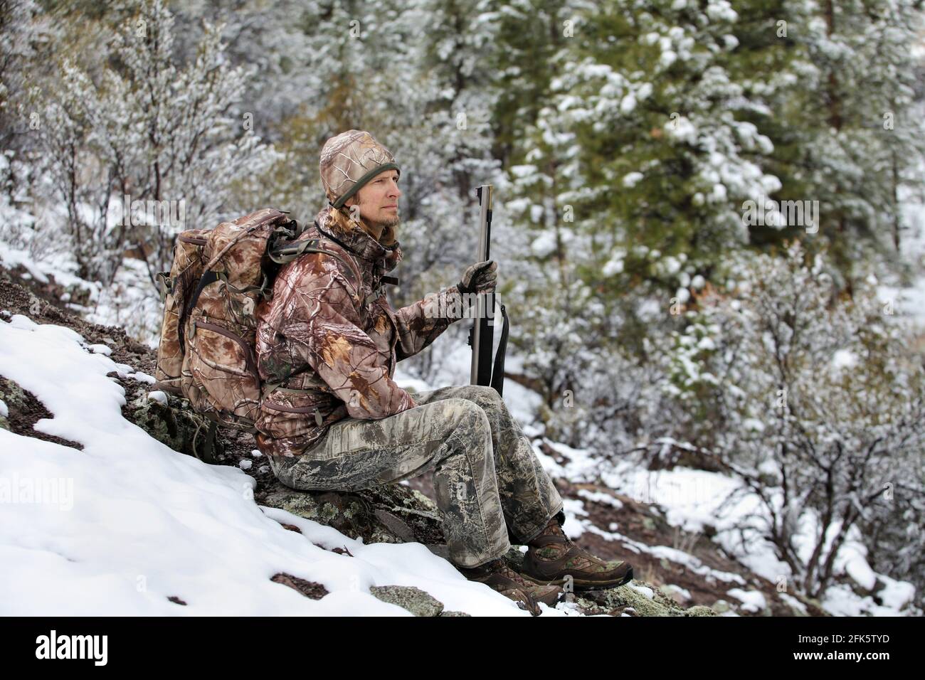 gun hunter wearing camouflage and sitting in snowy scene holding rifle Stock Photo