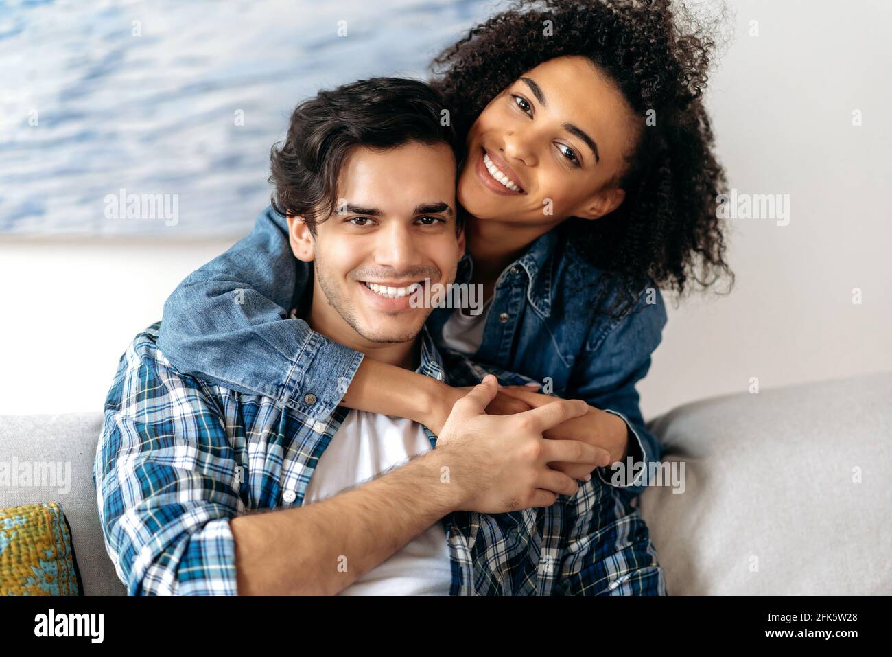 Portrait joyful happy mixed race couple, pretty african american curly girl tenderly hugging her hispanic boyfriend sitting on sofa, wearing casual clothes in living room, looking at camera, smiling Stock Photo