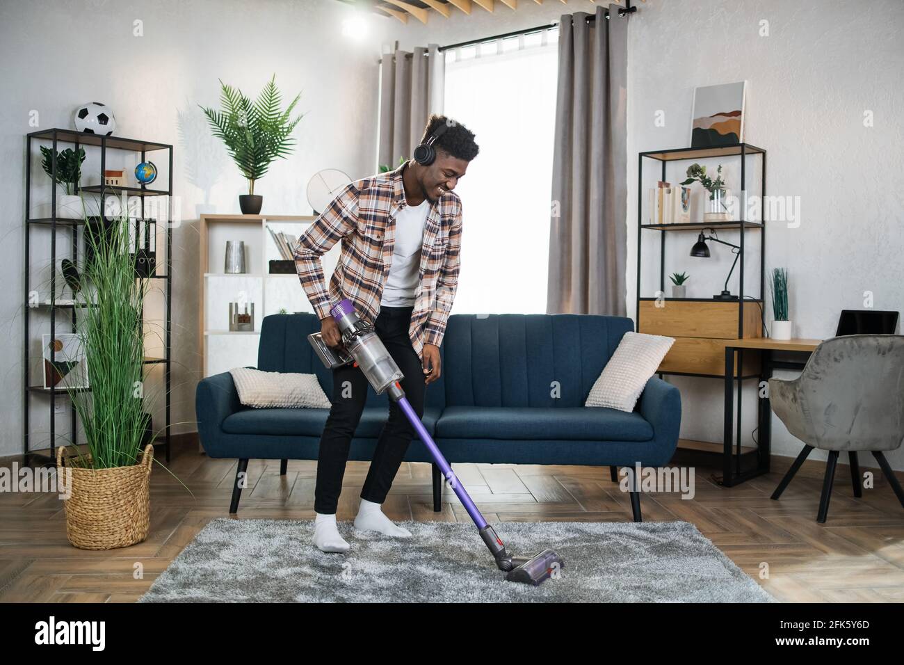 Smiling afro american man in wireless headphones cleaning carpet with  handheld vacuum cleaner. Young guy enjoying housekeeping using modern  devices Stock Photo - Alamy