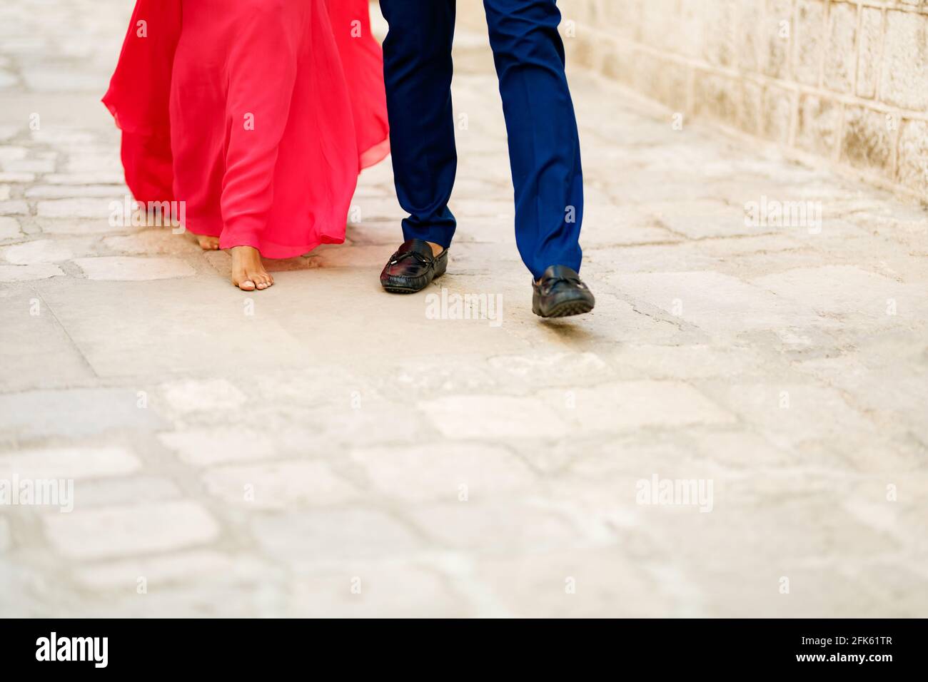 Barefoot bride in a long bright pink dress and the groom walking along a cobbled road, close-up Stock Photo