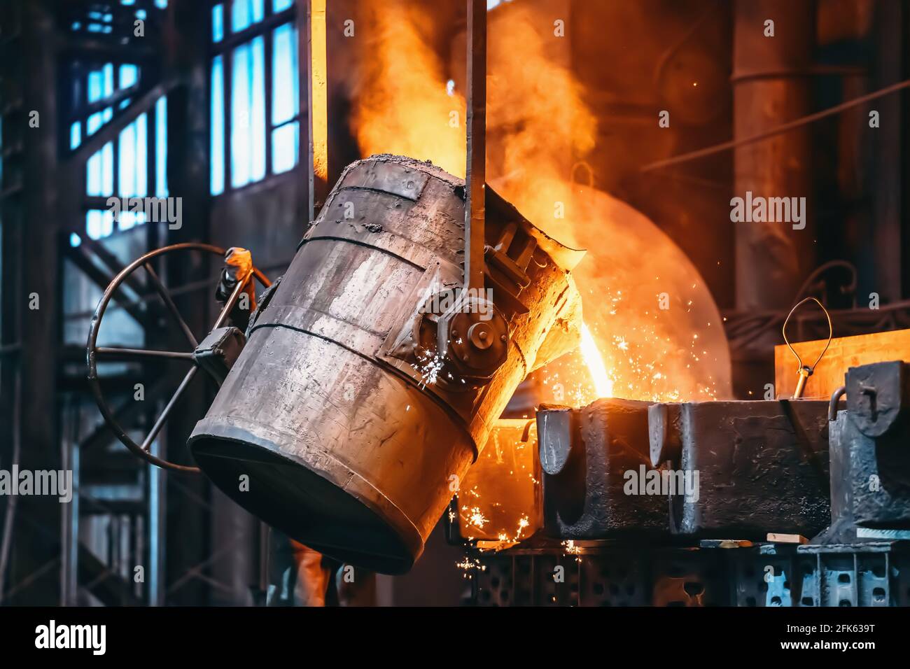 Metal casting process in foundry, liquid metal pouring from container to mold with clubs of steam and sparks, heavy metallurgy industry background. Stock Photo