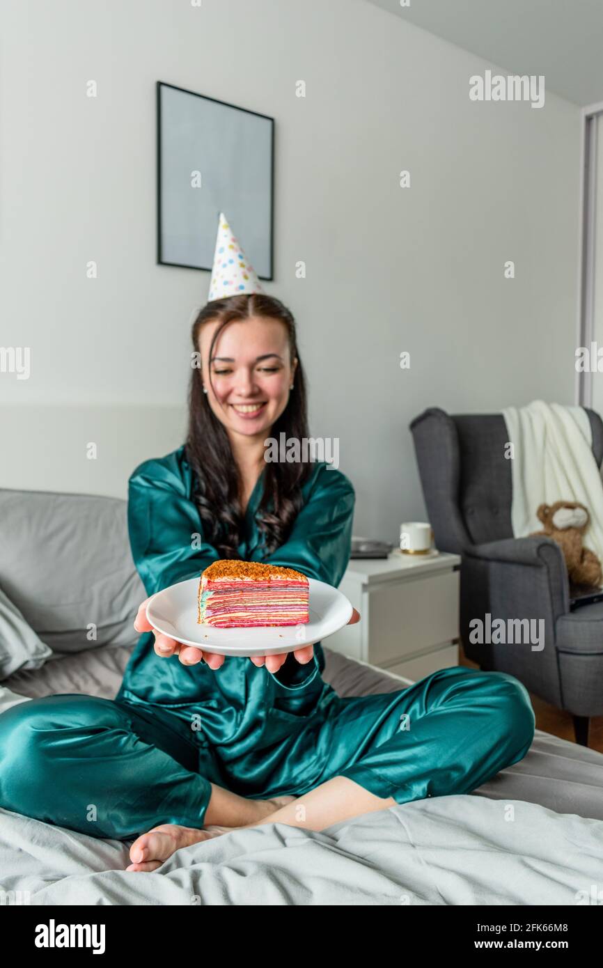 Young woman sits on the bed her pajamas and holds out cake on a plate. Wellness concept. holiday stay at home Stock Photo