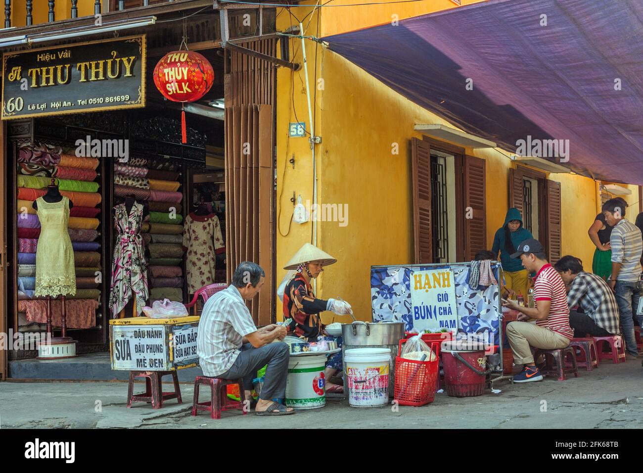 Vietnamese female selling street food at roadside cafe next to fabric shop in Old Town, Hoi An, Vietnam Stock Photo