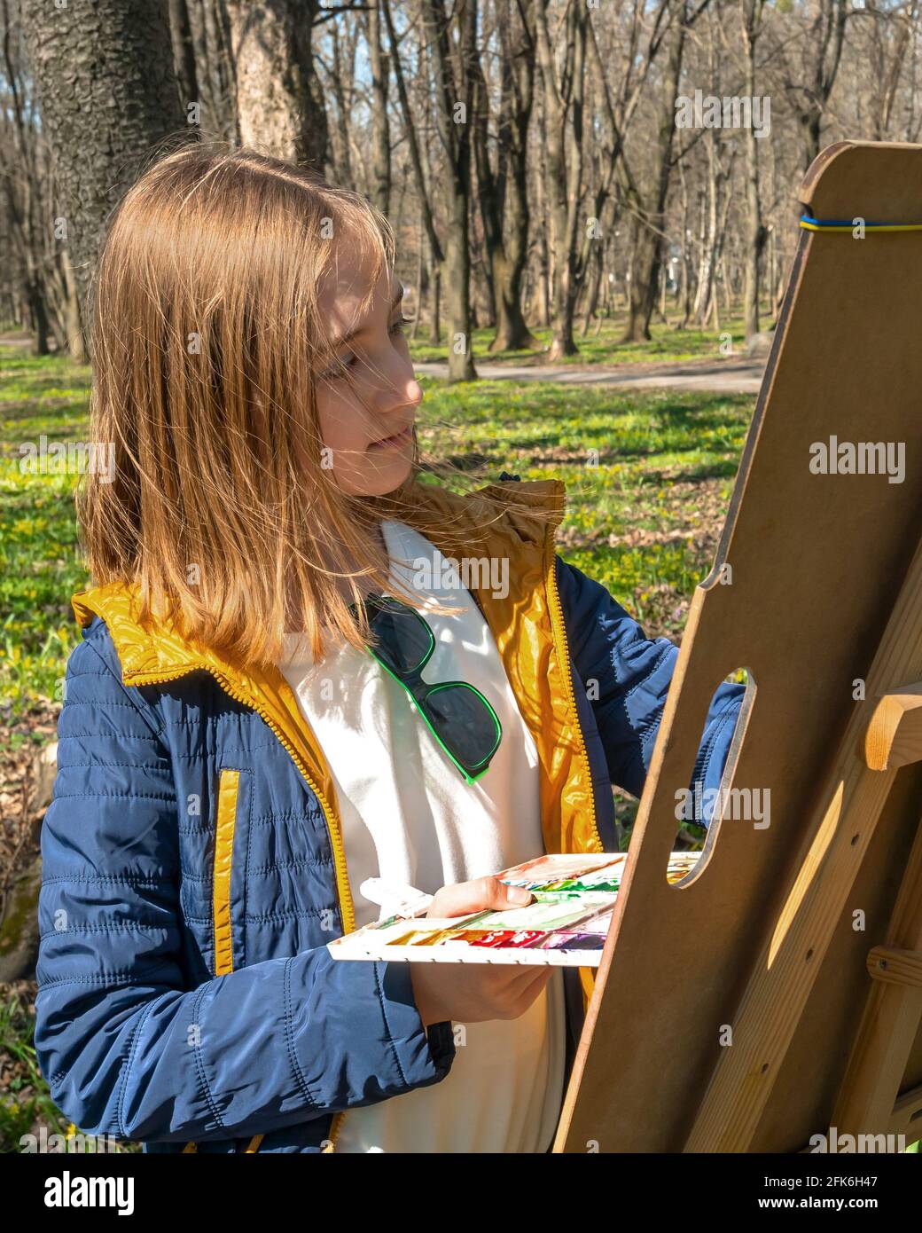 Beautiful Caucasian young girl with a palette in hand painted a picture on an easel. Park with trees and flowering plants in springtime, warm sunny we Stock Photo