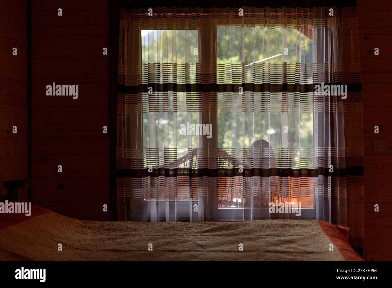 Relaxation and rest. Large window by the bed in the guest room of the wooden cottage. Girl is sitting on the terrace outside the window and enjoying h Stock Photo