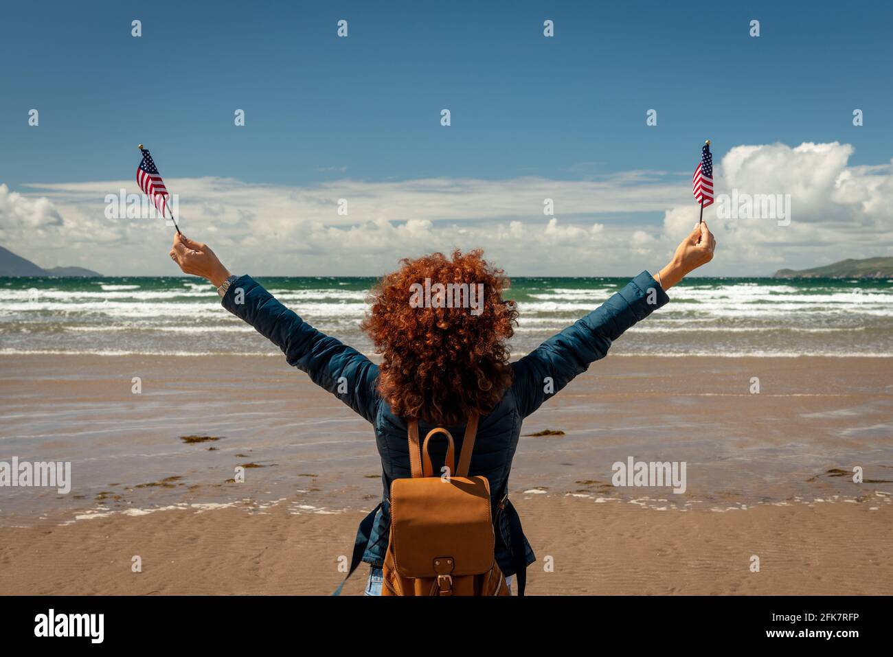 Rear view of young woman with red curly hair facing the sea while holding up American flags on a sandy beach on sunny day. Endless summer concept. Stock Photo