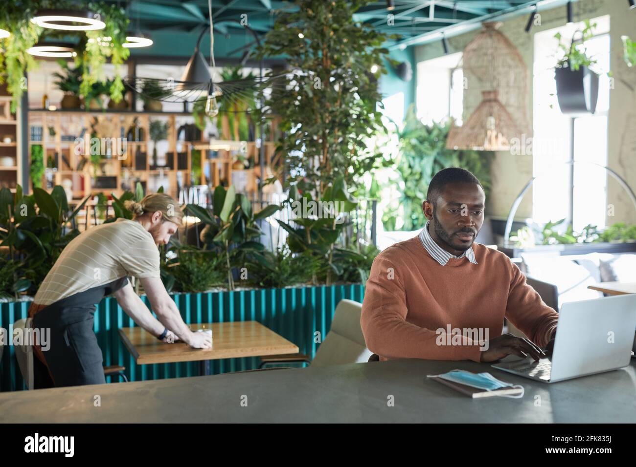 Portrait of African-American businessman using laptop while working in eco friendly cafe interior decorated with fresh green plants, copy space Stock Photo