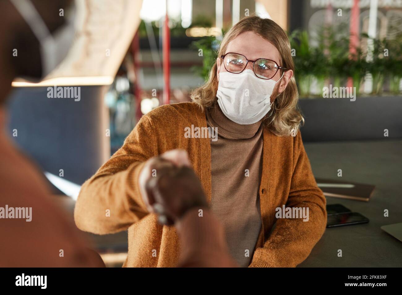Portrait of long haired man wearing mask and bumping fist with colleague during con tactless greeting in cafe or office, covid concept Stock Photo