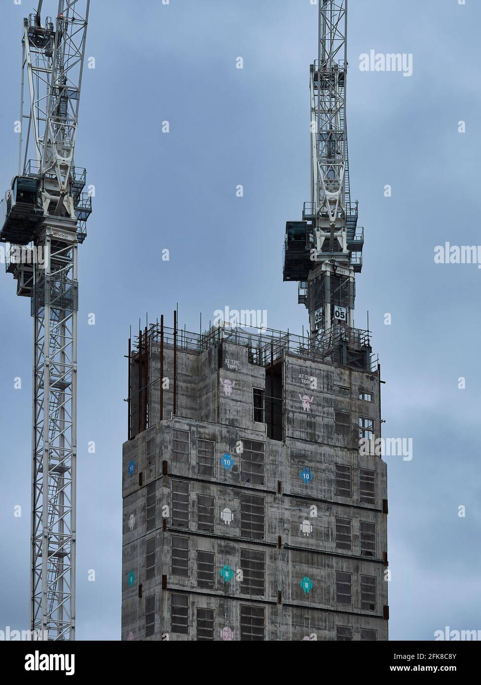 A pair of tower cranes, working to construct a central building core bearing Android logos, against an overcast grey sky. Stock Photo