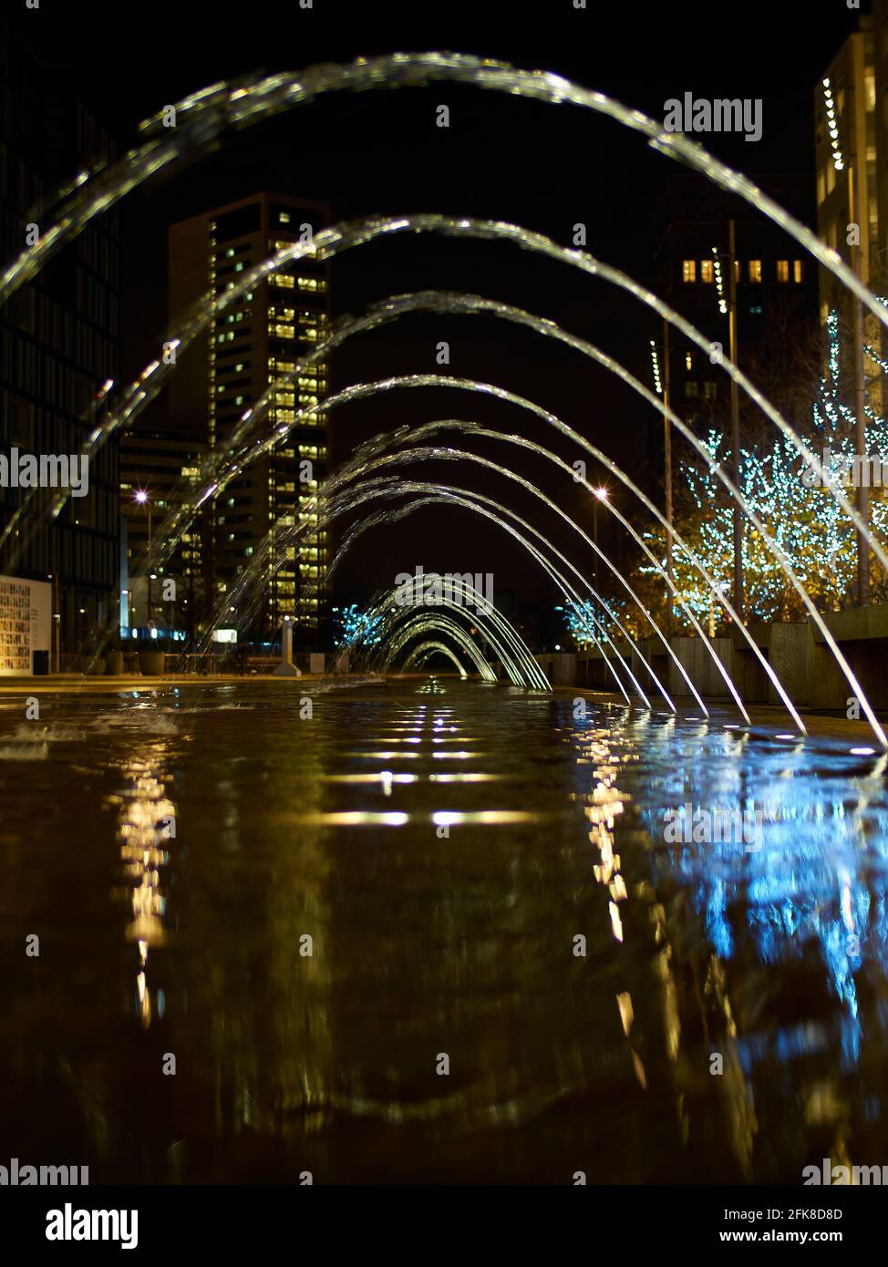 A slow-shutter, low angle view along a set of fountains, forming a tunnel with the blurred motion, surrounded by aspirational residential buildings. Stock Photo