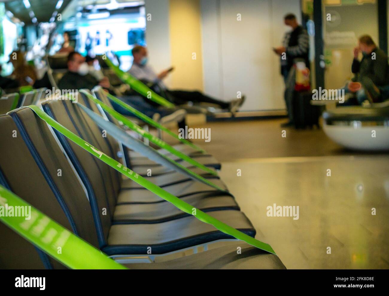 A row of black empty seats. Passengers in the transit zone of an airport, waiting in the sitting area, with distancing marked by green tape. Stock Photo