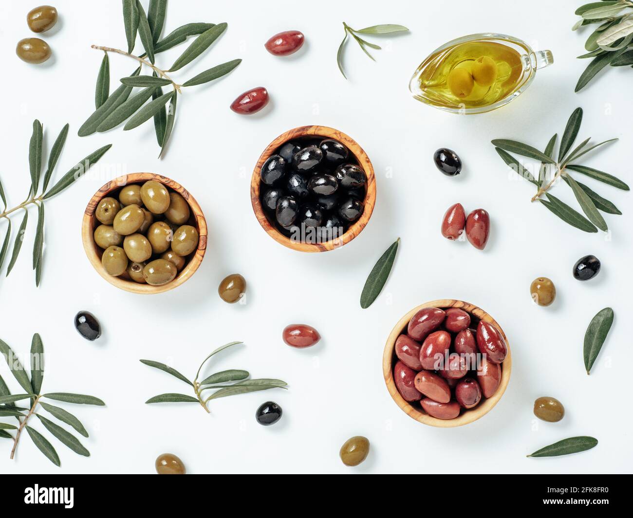 Set of green olives, black olives and red kalmata olives and extra virgin olive oil on white background. Top view of different types of olives in bowls and oil on leaves and branches background. Stock Photo