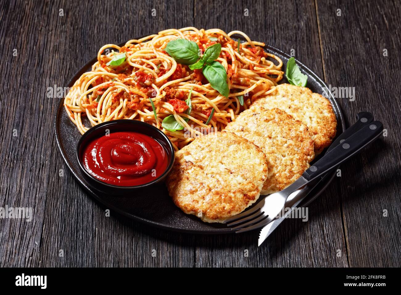 baked cod fish patties served on a black plate with spaghetti mixed with lentils, tomato sauce and herbs, horizontal view from above Stock Photo