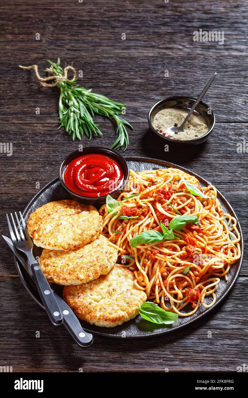 baked cod fish patties served on a black plate with spaghetti mixed with lentils, tomato sauce and herbs, vertical view from above Stock Photo