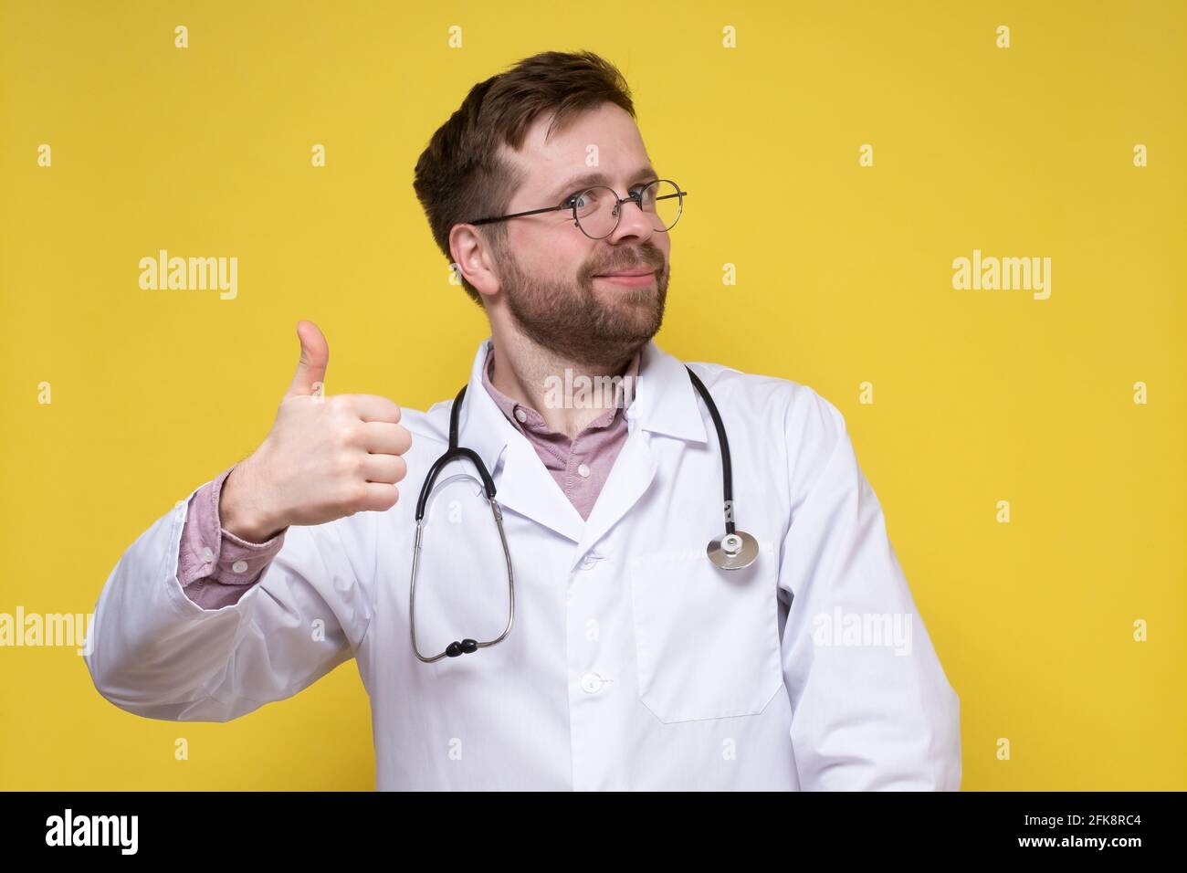 Cute, contented doctor shows an approving gesture, thumbs up and smiles friendly while looking into the camera.  Stock Photo