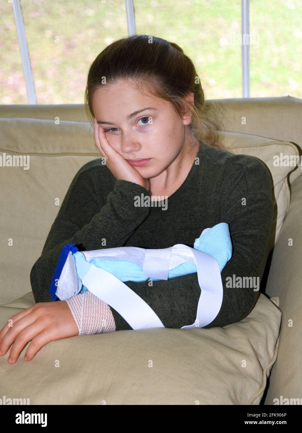 Little girl sits, very discouraged, with ice pack on her arm.  Injury is wrapped with bandage and is resting on a pillow inside her home.  She is rest Stock Photo
