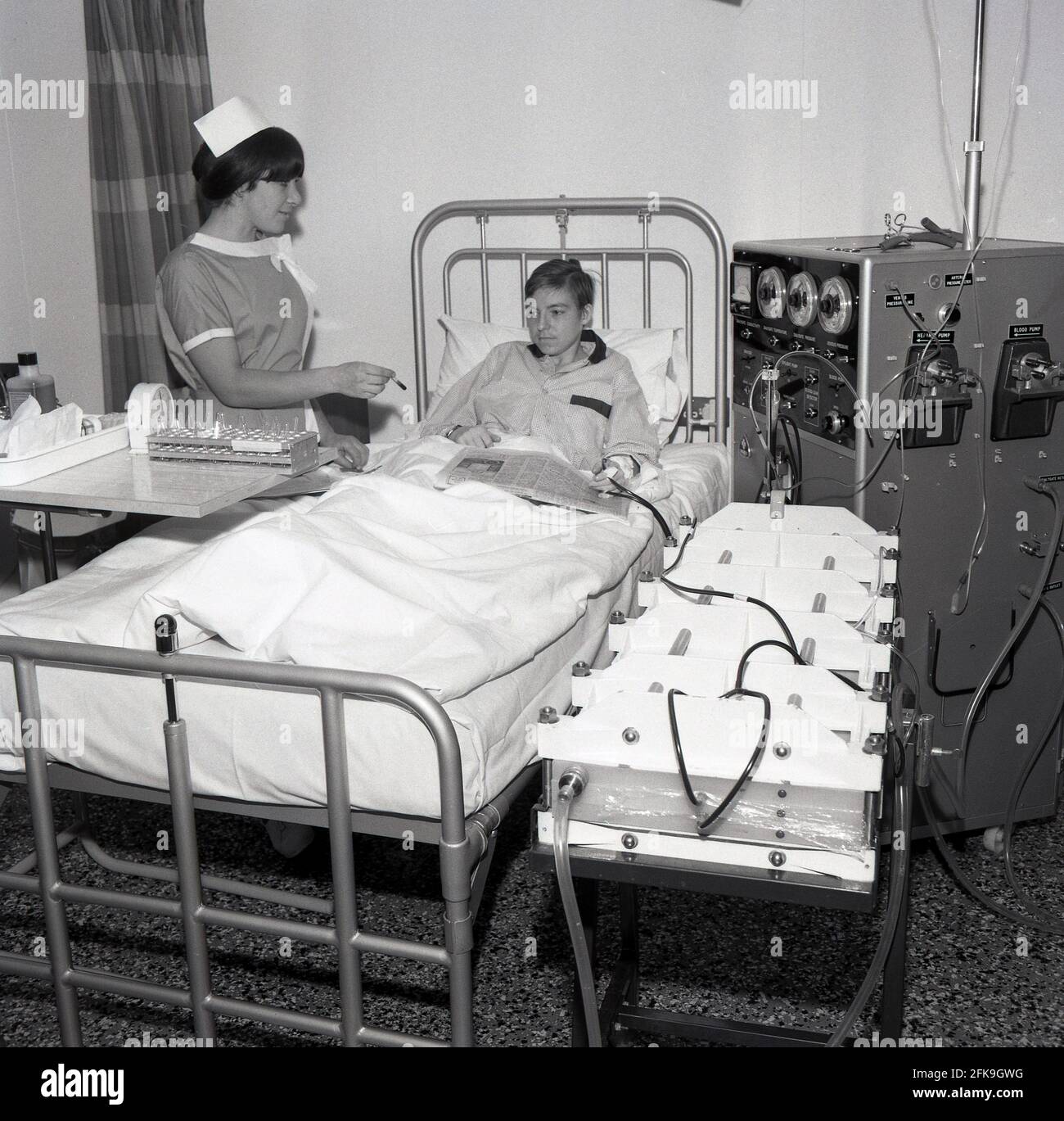 1960s, historical, a female nurse with a small test tube, standing beside a young lad lying in a hospital bed with a broadsheet newspaper on his lap. He is linked up to a large machine, a dialysis machine that cleans blood through a filter known as a dialyzer, South East London, England, UK. Dialysis is a medical treatment that filters and cleans the blood using a machine, which helps to keep the body in balance when the kidneys can't perform this function. Stock Photo