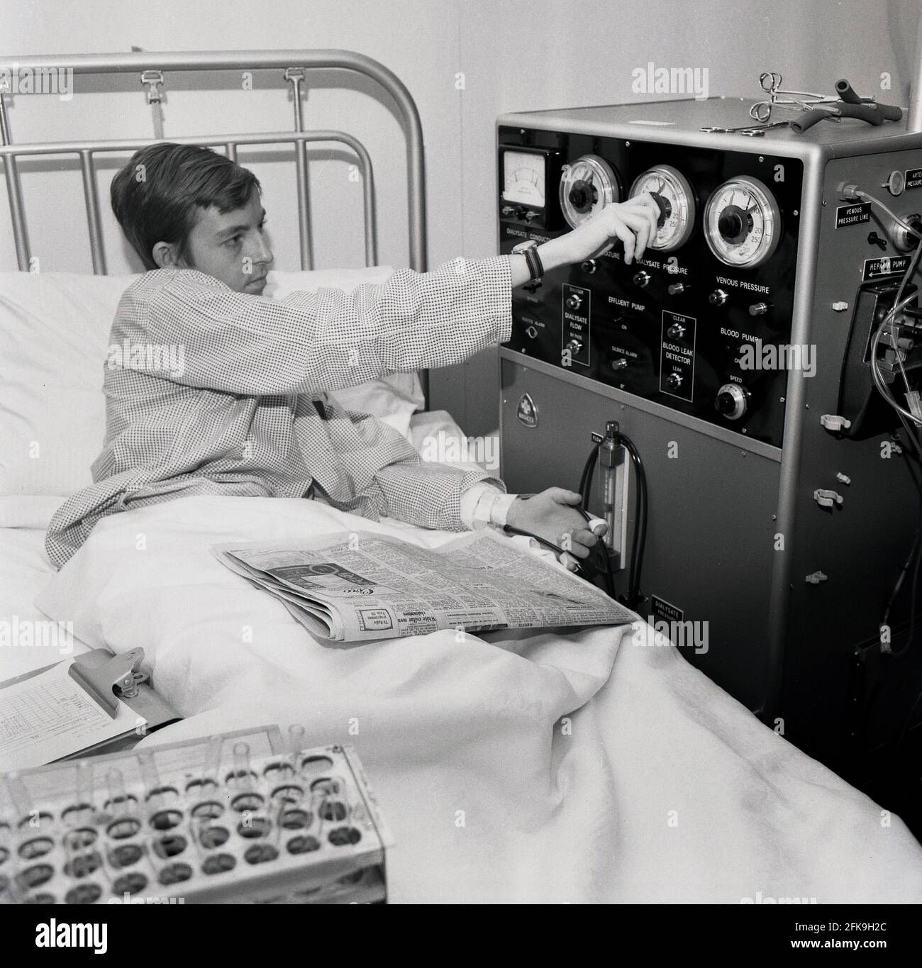 1960s, historical, a young man lying in a hospital bed adjusting a dial on a large machine he is attached to, a dialaysis machine that helps pump blood into the kidneys, South East London, England, UK. Dialysis is a medical treatment that filters and cleans the blood using a machine, which helps to keep the body in balance when the kidneys can't perform this function. Also known as Hemodialysis Machines. HD machines clean blood by passing it through a filter called a dialyzer. Stock Photo