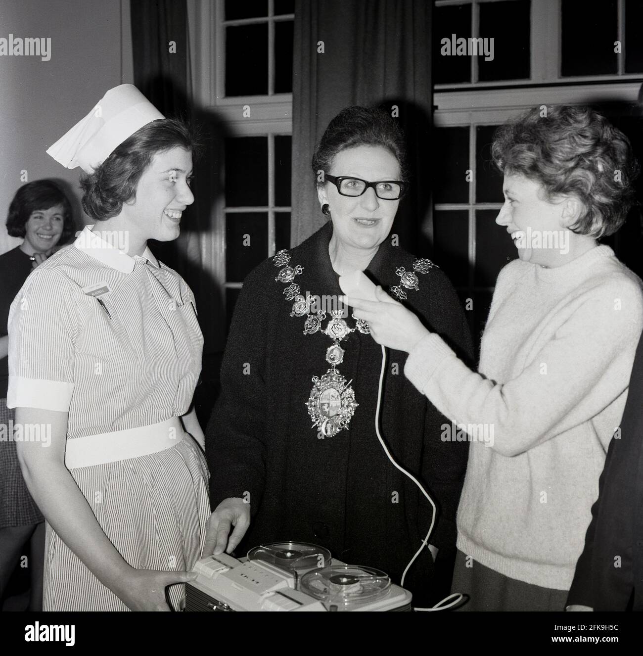 1960s, historical, at a South East London hospital, a nurse and local female journalist stand beside a Lady Mayores, recording her speaking with a portable reel to reel tape recorder. Stock Photo