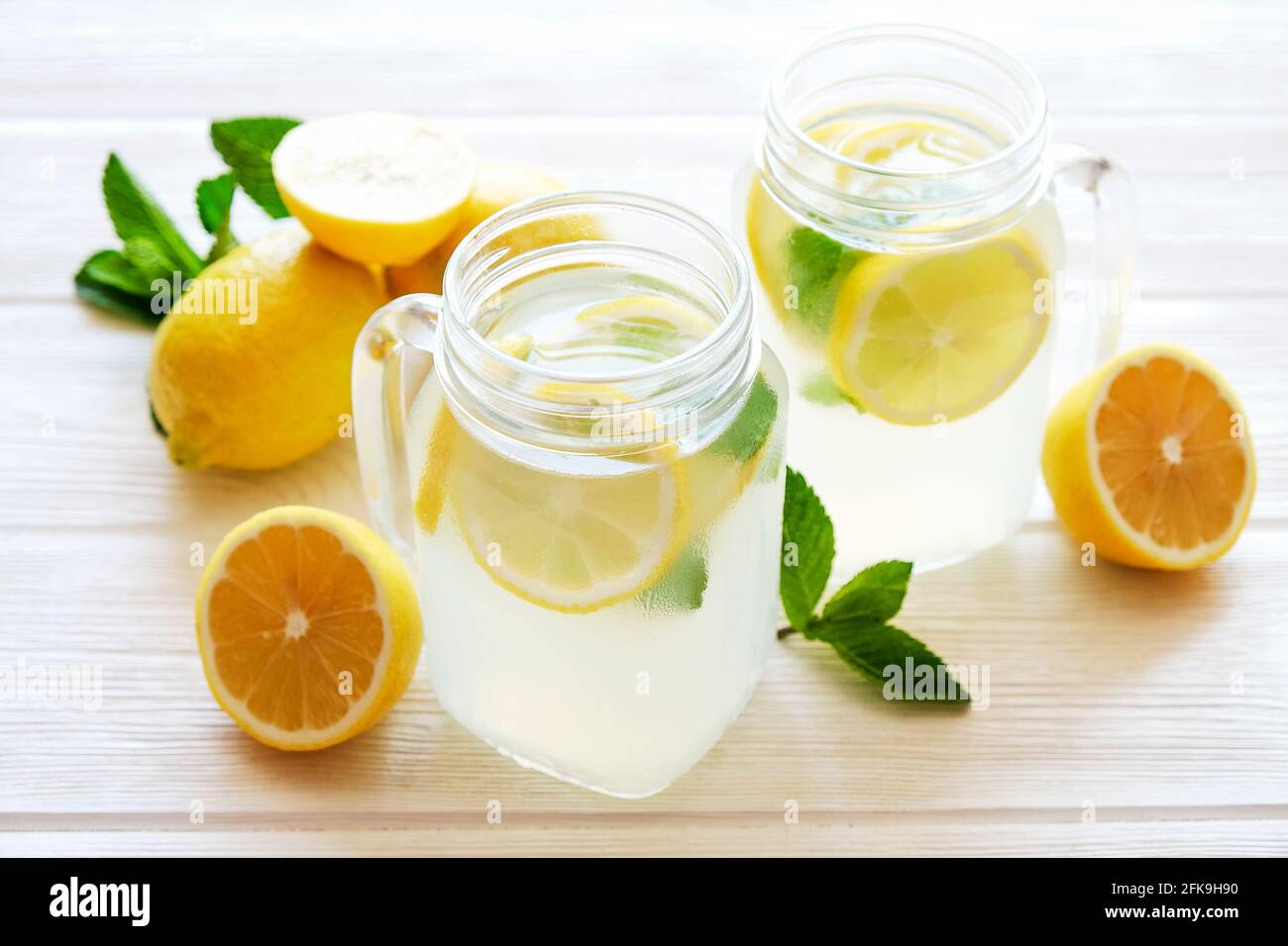 Two mason jar glasses of homemade refreshing lemonade with slices of organic ripe lemon, whole and halved. Non alcoholic beverage on a rustic white wo Stock Photo