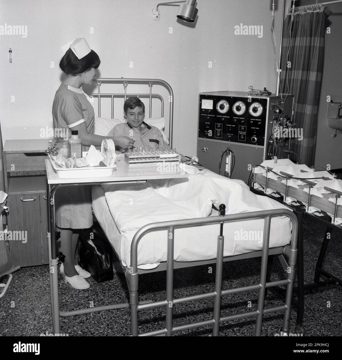 1960s, historical, a female nurse with test tubes of blood samples standing beside a young man lying in a hospital bed linked up to a large machine, a kidney dialaysis machine, South East London, England, UK. Dialysis is a medical treatment that filters and cleans the blood using a machine, which helps to keep the body in balance when the kidneys can't perform this function. Also known as Hemodialysis Machines. HD machines clean blood by passing it through a filter called a dialyzer. Stock Photo