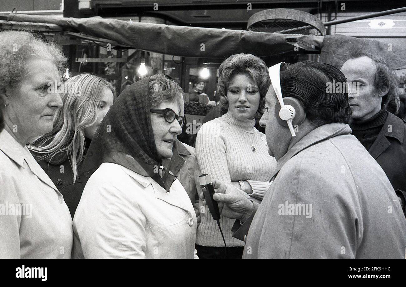 1970s, popular man of the people, radio presenter and reporter, Monty Modlyn recording a local woman's opinion, with other people listening in, outside in a South London market area, England, UK. Born into a Jewish family from Lambeth, Modlyn was known for his cockney accent and his broadcast forte was interviewing ordinary people, a technique that became known as 'Vox Pop'. Addressing people with the words 'Ullo darlin', his east end working class persona saw him have conversations about anything and everything during a long and distinguished broadcasting career. Stock Photo
