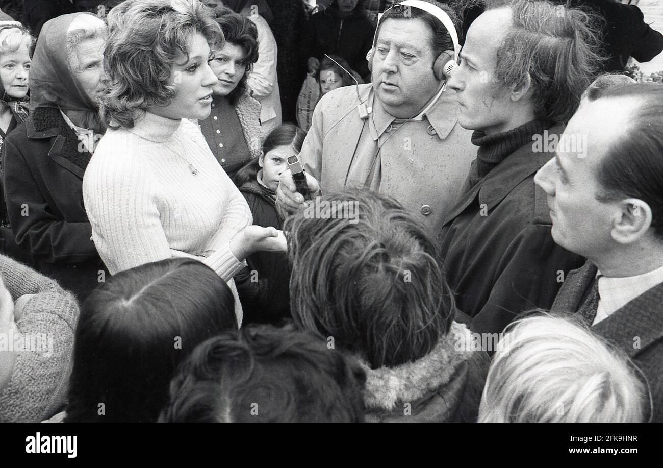 1970s, popular man of the people, radio presenter and reporter, Monty Modlyn with headphones and microphone lisiting to an attractive woman talking, surrounded by other people in a South London market area, England, UK. Born into a Jewish family from Lambeth, Modlyn was known for his cockney accent and his broadcast forte was interviewing ordinary people, a technique that became known as 'Vox Pop'. Addressing people with the words 'Ullo darlin', his east end working class persona saw him have conversations about anything and everything during a long and distinguished broadcasting career. Stock Photo