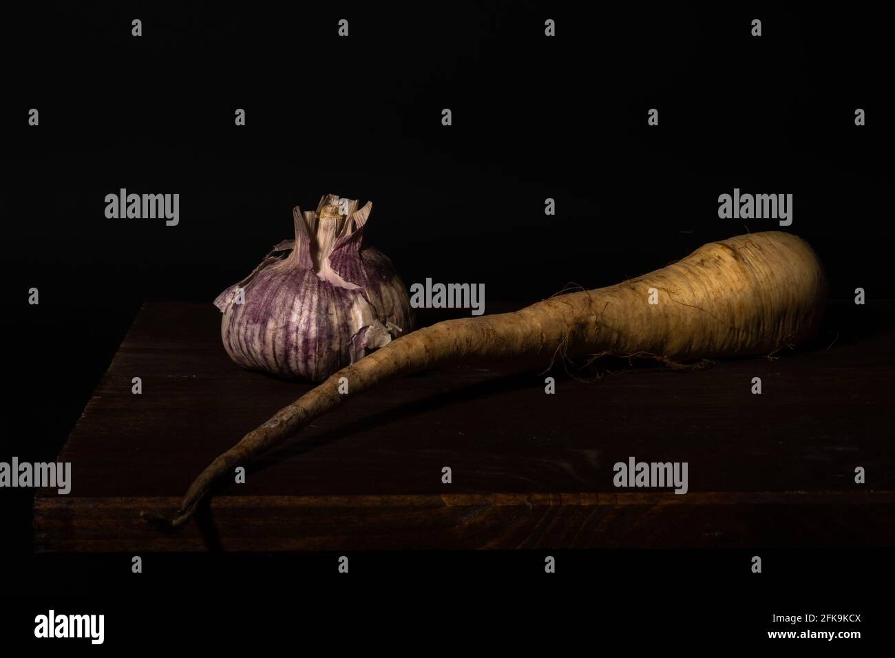 A Stillife of garlic and a parsnip on a wooden table against a dark background Stock Photo