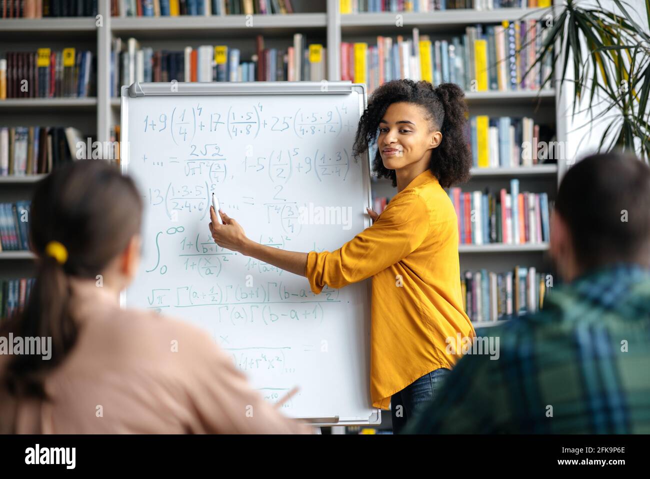 Attentive smart students listen to young mixed race woman teacher standing by whiteboard, wearing stylish clothes, is conducts lesson, brainstorm, shows information and smiling Stock Photo