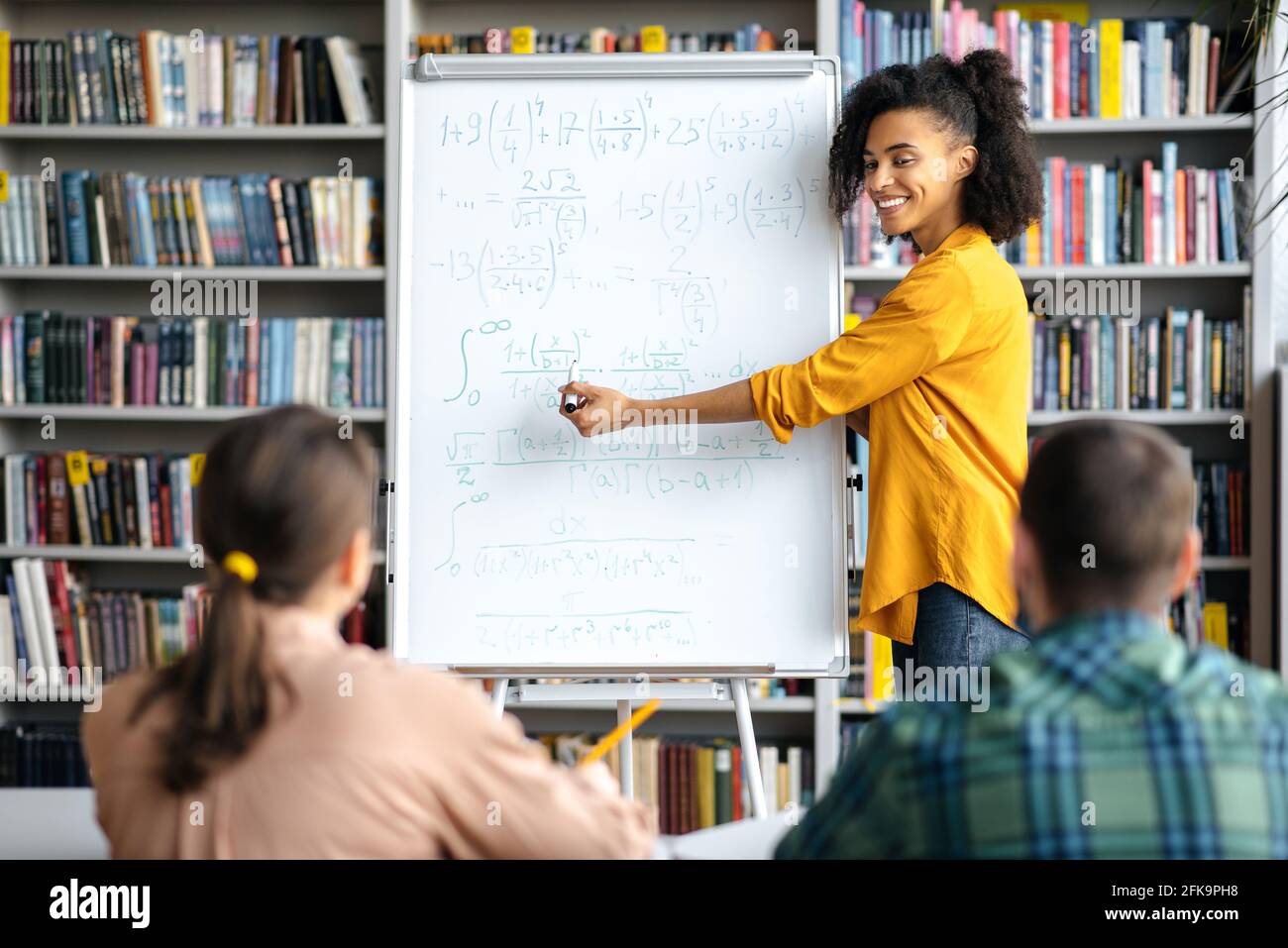 Attentive smart students listen to young mixed race woman teacher standing by whiteboard, wearing stylish clothes, is conducts lesson, brainstorm, shows information and smiling Stock Photo