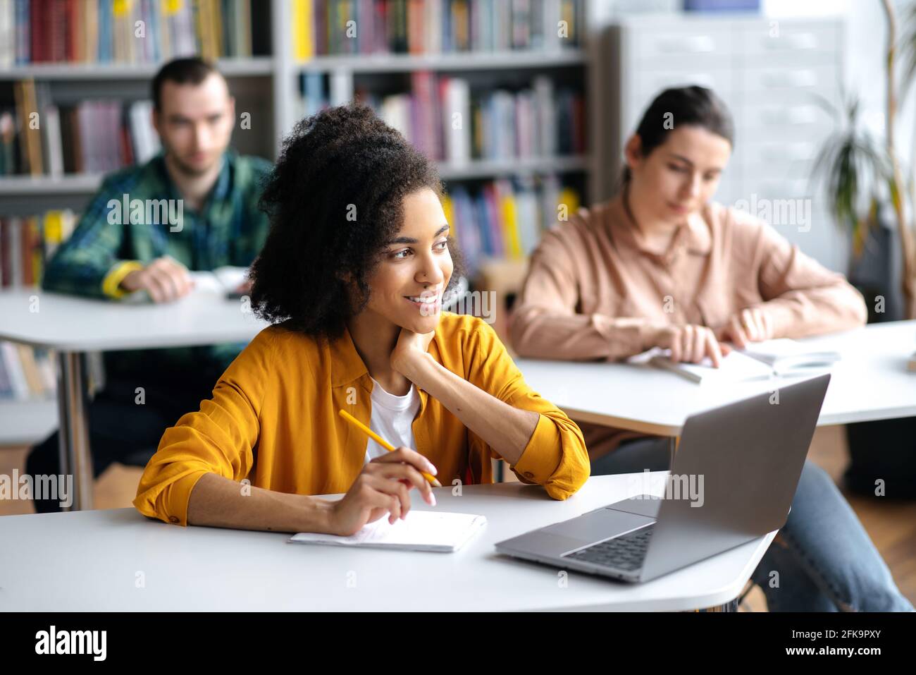 Joyful smart cute young african american female student, in casual informal clothes, sitting at table in university library with laptop while preparing for lessons or exam, taking notes, smiling Stock Photo
