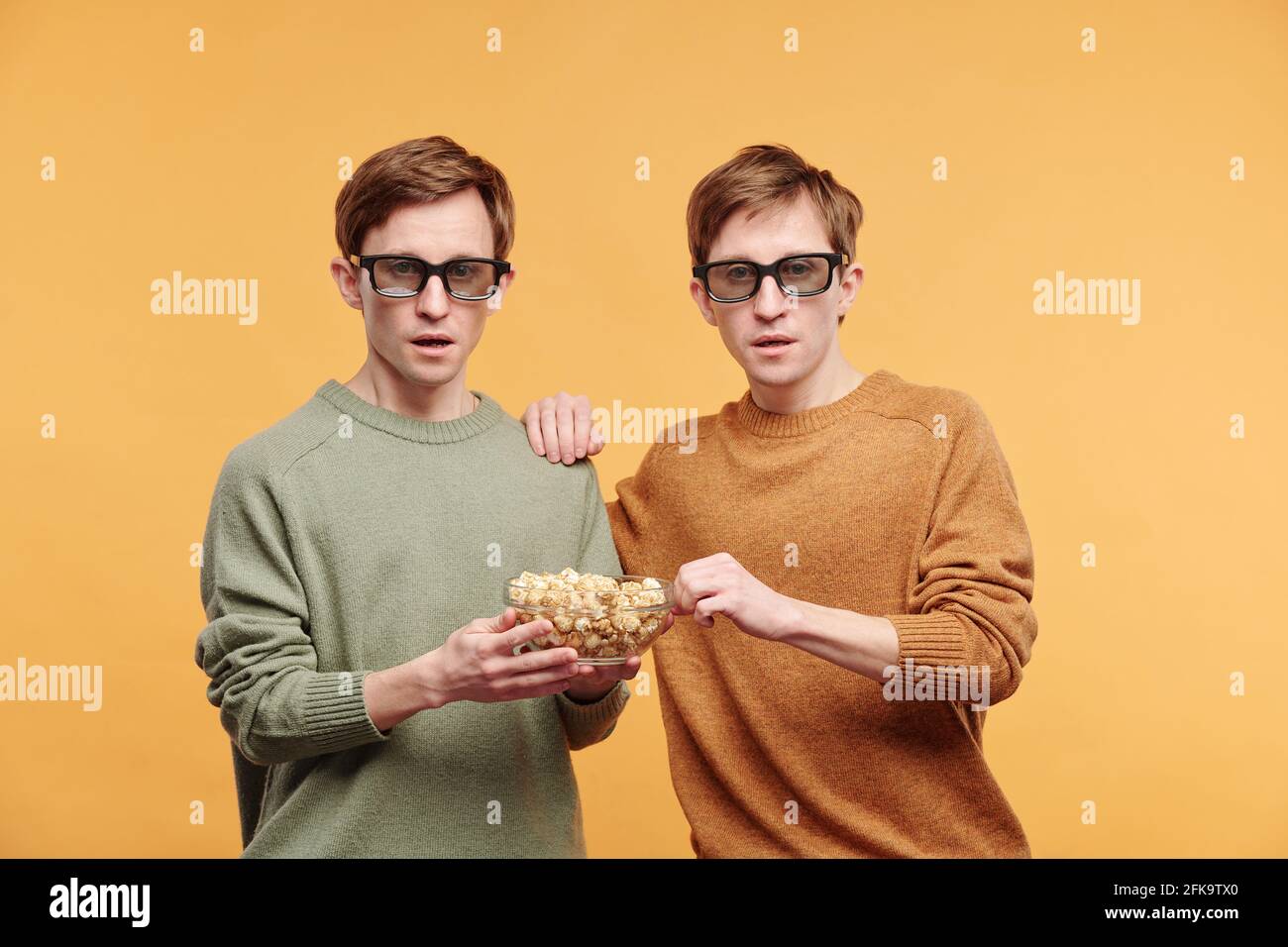 Portrait of shocked curious twins in sweaters and 3D goggles eating popcorn while watching movie against orange background Stock Photo