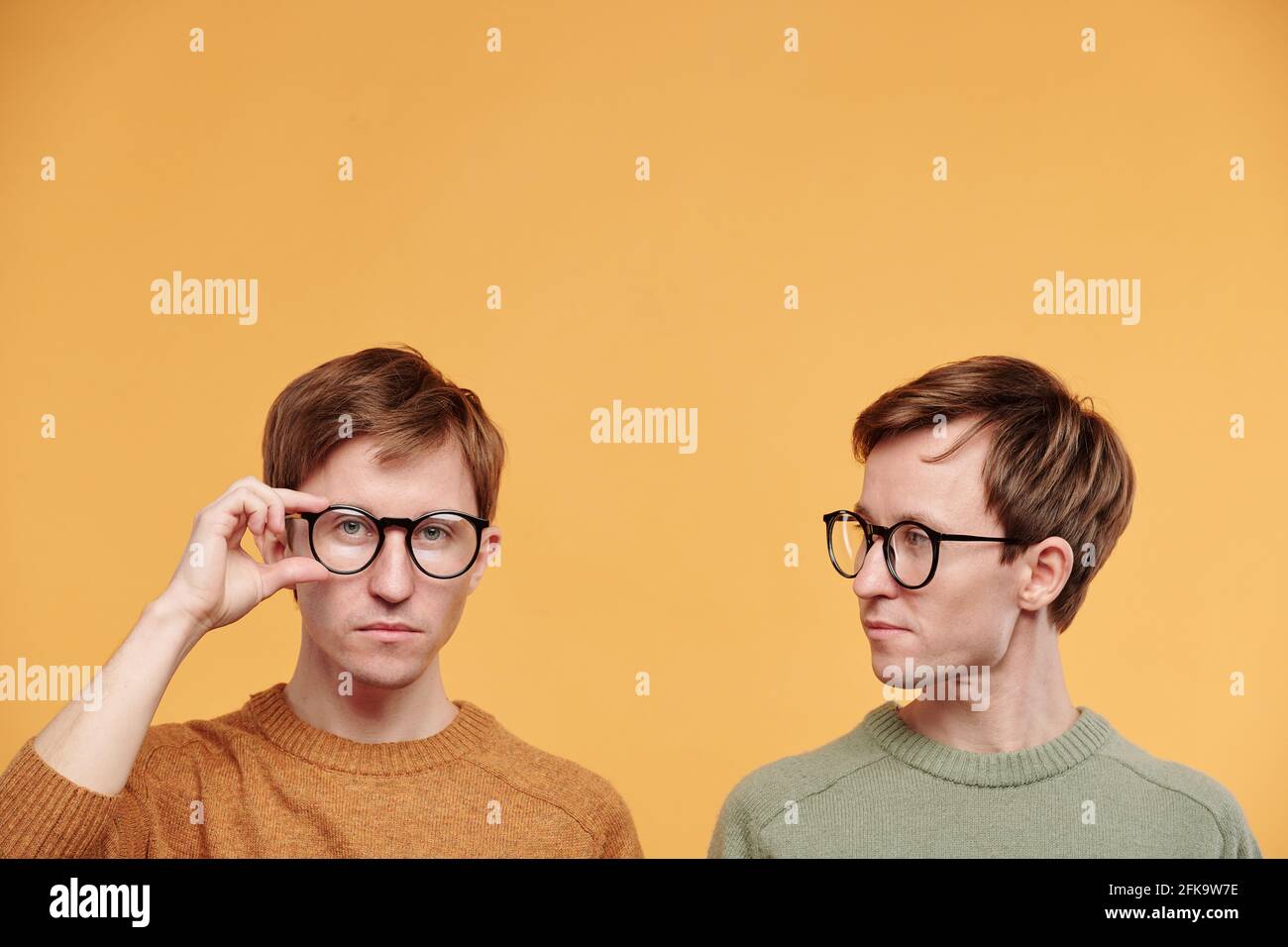 Young fair-haired man in olive sweater looking at brother adjusting glasses against orange background Stock Photo