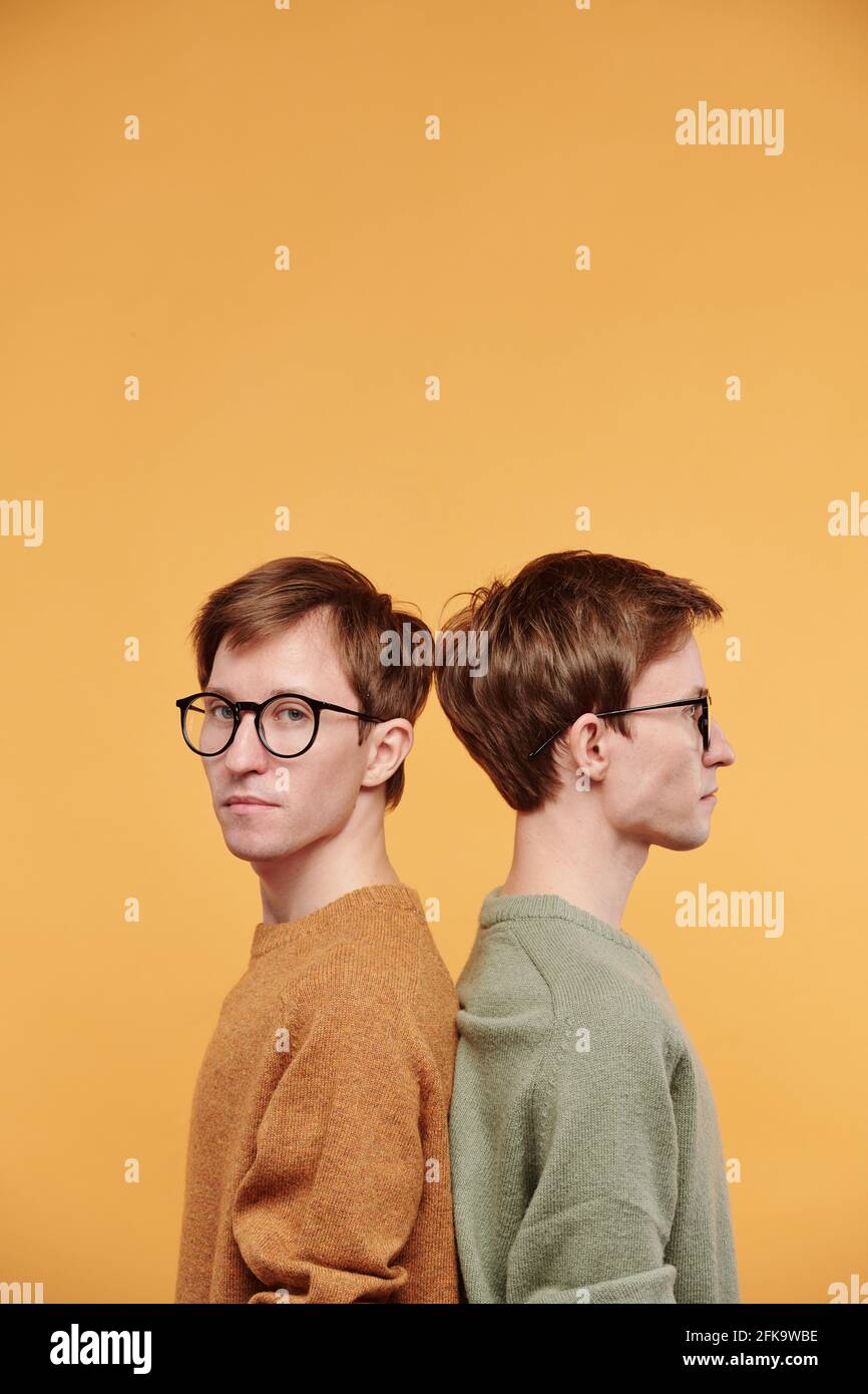 Portrait of serious handsome young man in glasses standing back to back with twin brother against bright background Stock Photo