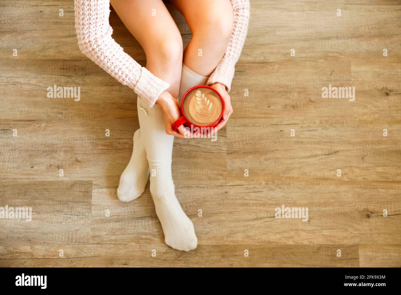 Young woman drinking cappuccino coffee and sitting on the wooden floor. Top view of female legs in warm white knee high socks. Comfort winter holidays Stock Photo